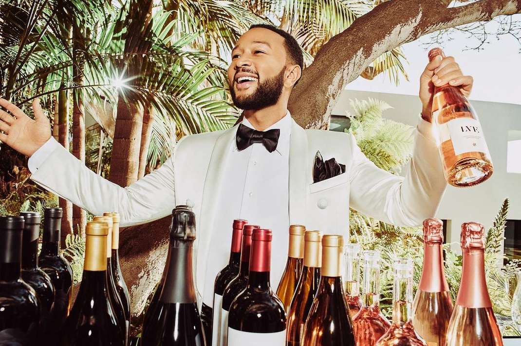 John Legend in a white tuxedo smiles and gestures, holding a rosé bottle. He stands behind a bar filled with various wine bottles.
