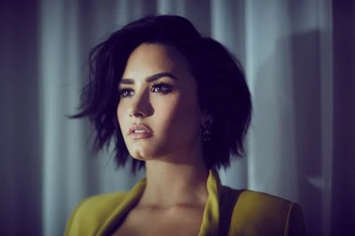Portrait of Demi Lovato with dark short hair wearing a mustard yellow jacket against a draped curtain background