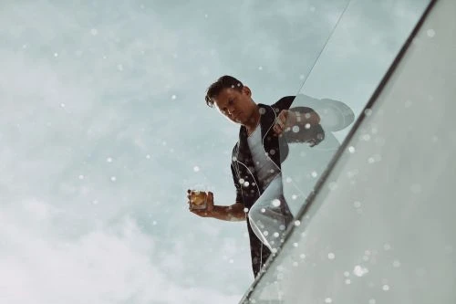 A man in a dark robe and white t-shirt holding a glass of whiskey while leaning on a glass balcony railing outdoors.