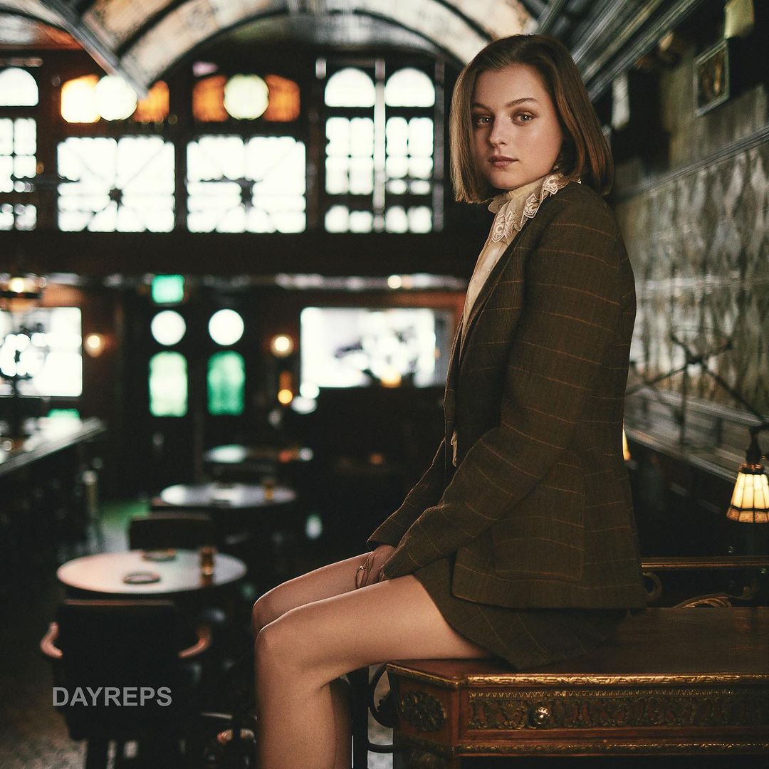 Indoor portrait of a woman with short brown hair, wearing a tailored brown jacket and skirt, seated on a decorative table in a bar.