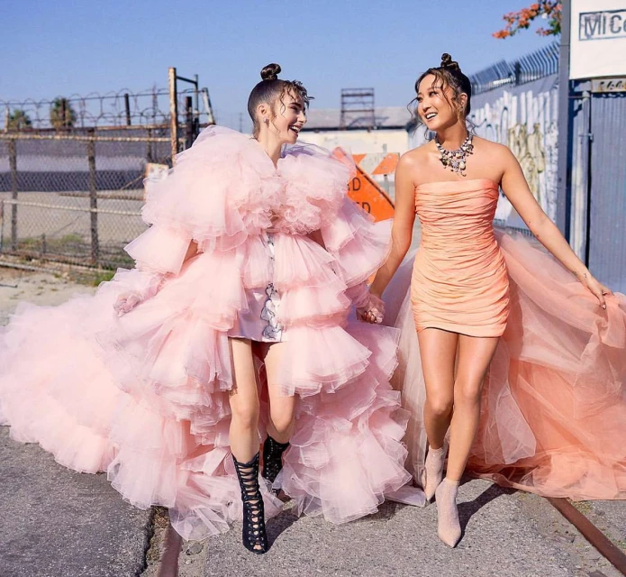Two women in formal dresses pose on a city street, one in a pink ruffled gown with train, the other in a peach ruched mini-dress.