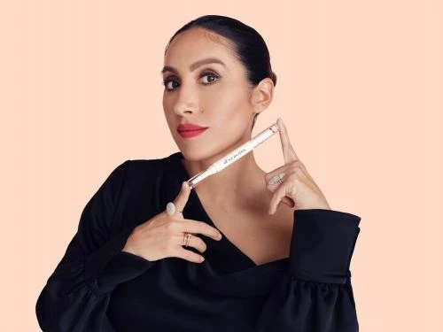 Portrait of an adult female holding a white cosmetic pen and wearing a black dress against a peach-colored background.