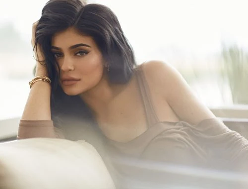 Portrait of a young adult female with dark hair in soft lighting, wearing a brown top and gold bracelet, with neutral makeup.