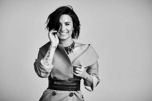 A woman with short dark hair smiles in a black and white studio portrait, wearing a belted coat and necklace, with a tattoo on her arm.