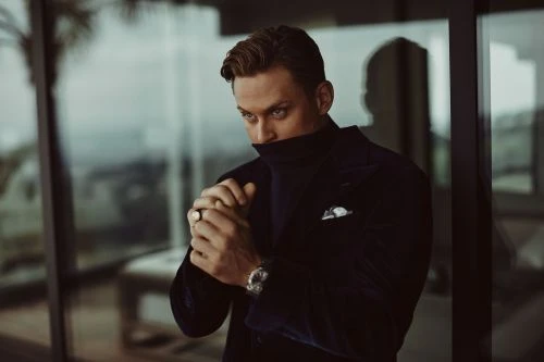 Portrait of a man in a dark blue velvet suit, his hands clasped near his face, standing indoors near a window with a blurred background.