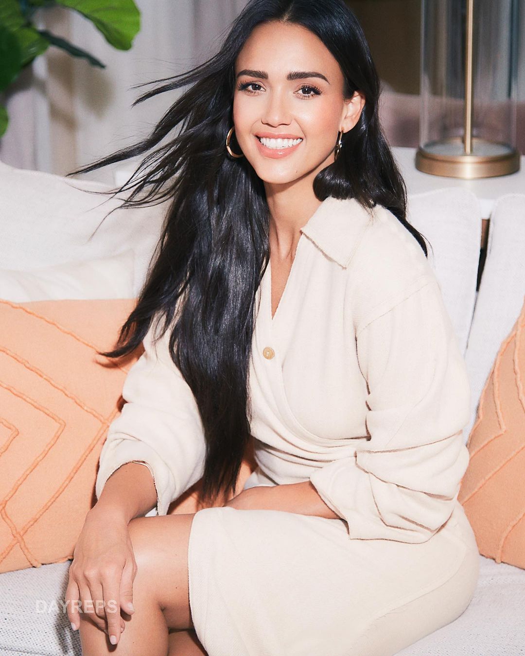 Smiling woman with long dark hair sitting on a couch with decorative pillows in a light-filled interior space wearing a cream-colored dress.