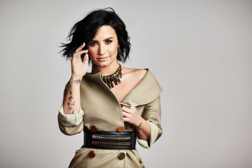 Elegant portrait of a woman with dark hair in a draped tan jacket, accessorized with a bold necklace and wide belt against a light backdrop.