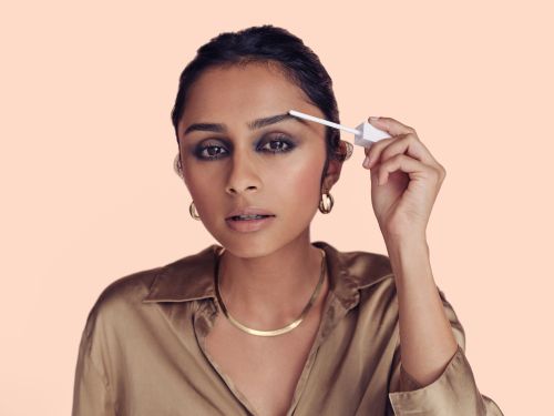 Adult woman applying brow gel with a wand. She is wearing a tan collared shirt, gold jewelry, and dark eye makeup, with light peach background.