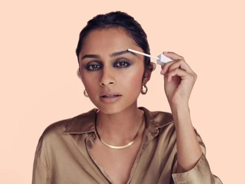 Adult woman applying brow gel with a wand. She is wearing a tan collared shirt, gold jewelry, and dark eye makeup, with light peach background.