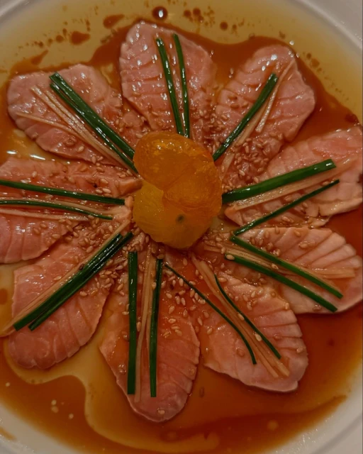 Overhead shot of salmon sashimi arranged like a flower, with green onions and sesame seeds, drizzled with sauce and garnished with a yellow fruit.