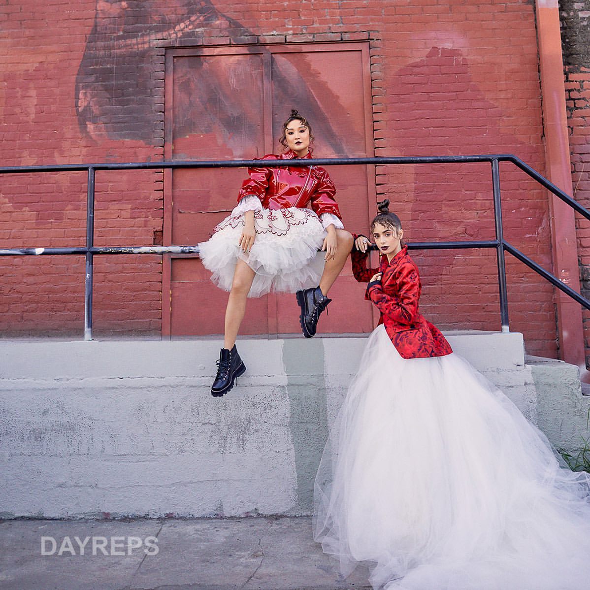 Two models pose in tutus and red jackets on concrete steps with a brick wall backdrop, showcasing a blend of fashion and urban style.