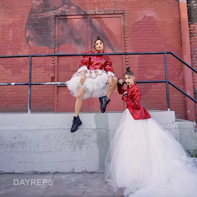 Two models pose in tutus and red jackets on concrete steps with a brick wall backdrop, showcasing a blend of fashion and urban style.