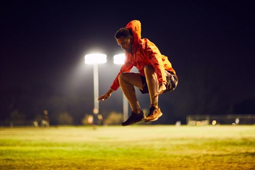 Athletic man jumping in mid-air on grassy sports field at night, wearing an orange hoodie, black shorts, and running shoes.