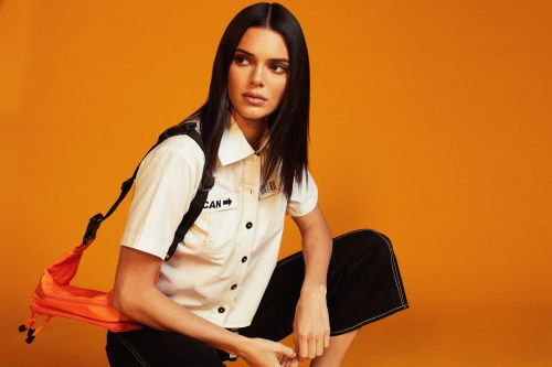 Woman with dark hair poses in a white button-down shirt with a black backpack strap and orange bag, set against a bright orange background.