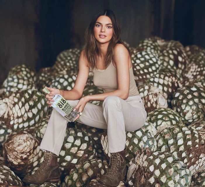Adult female model sits on pile of agave plants in desert environment holding bottle of 818 Tequila. Light skin, brown hair, neutral colours.