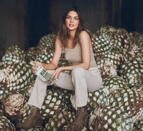 Adult female model sits on pile of agave plants in desert environment holding bottle of 818 Tequila. Light skin, brown hair, neutral colours.