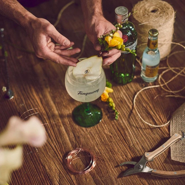 Overhead shot of a mixologist's hands garnishing a cocktail with a pear slice and flowers on a rustic wooden table with bar tools.