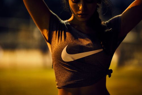 Woman in athletic shirt with Nike logo with arms raised poses outdoors at night with diffuse lighting and a shallow depth of field.