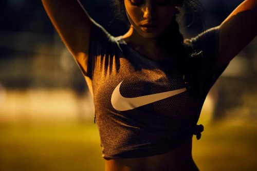 Woman in athletic shirt with Nike logo with arms raised poses outdoors at night with diffuse lighting and a shallow depth of field.