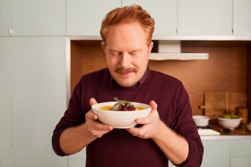 Red-haired adult male holding a bowl of food in a kitchen, eyes closed, face tilted to savor the aroma.