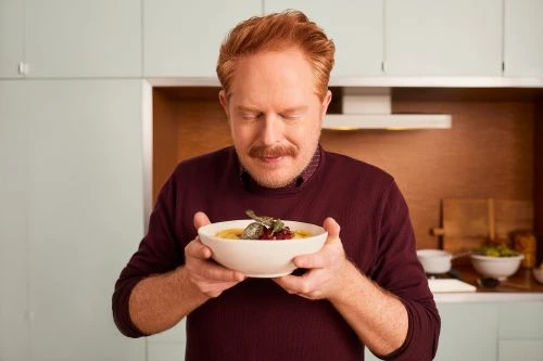 Red-haired adult male holding a bowl of food in a kitchen, eyes closed, face tilted to savor the aroma.