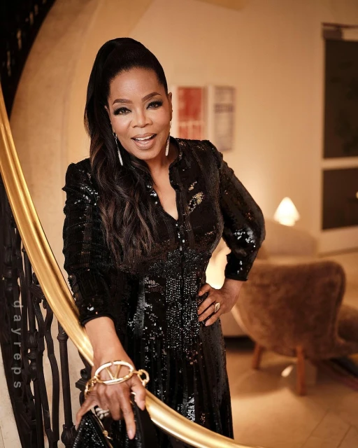 Oprah Winfrey poses in a black, sequined dress on a golden staircase indoors, with elegant decor in the background. Portrait shot.