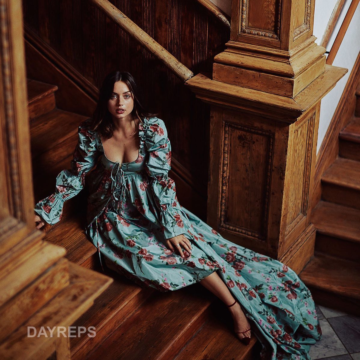 Woman in a floral dress sits on a wooden staircase in an indoor setting, illuminated by soft, diffused lighting, creating an elegant portrait.