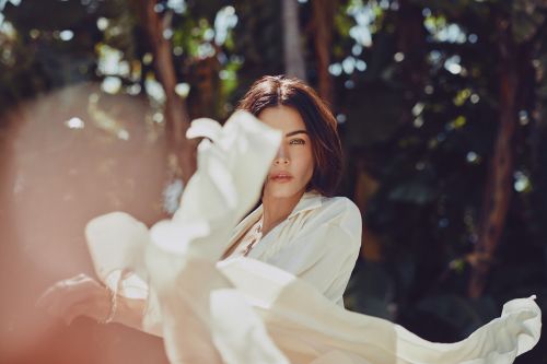 Portrait of a woman with long hair wearing a light shirt and holding a white textile outdoors in a forest-like setting with bokeh.