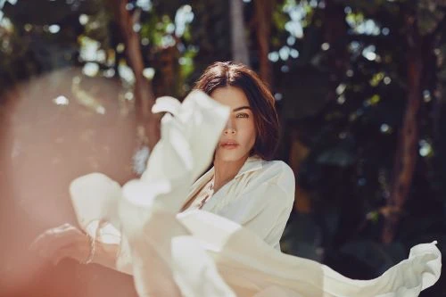 Portrait of a woman with long hair wearing a light shirt and holding a white textile outdoors in a forest-like setting with bokeh.