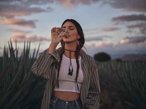 Woman drinks from glass outdoors at dusk with a field of agave plants. She wears a striped shirt, white top, jeans and choker.