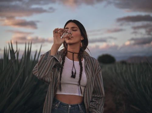 Woman drinks from glass outdoors at dusk with a field of agave plants. She wears a striped shirt, white top, jeans and choker.