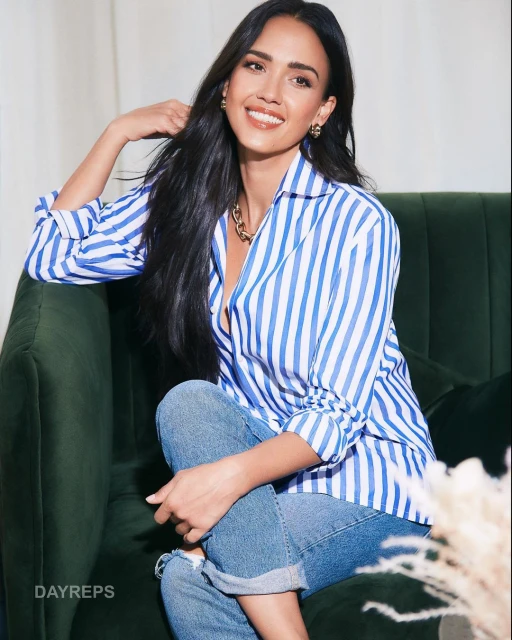 Portrait of a smiling adult woman with long dark hair and blue jeans sitting on a green velvet couch in a light-filled environment.