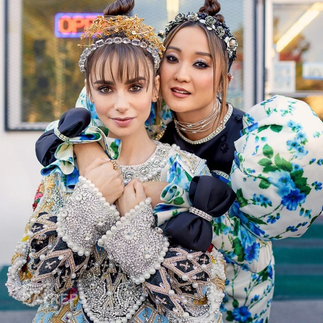 Two adult women pose outdoors, wearing elaborate floral and pearl-embellished dresses and jeweled headbands, with a shopfront in the background.