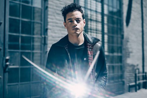 Portrait of actor Rami Malek outdoors in a black leather jacket and a plaid shirt, standing against a concrete building with glass block windows.