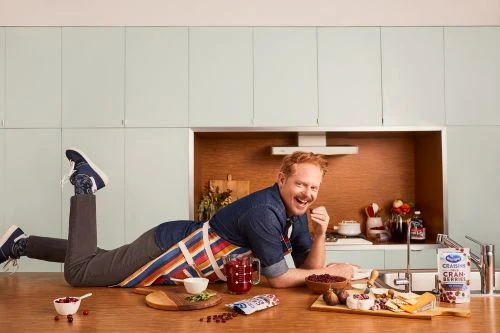 Smiling adult male lies on a kitchen counter with cranberries, cheeses, fruit, and a drink in front of light green cabinets.