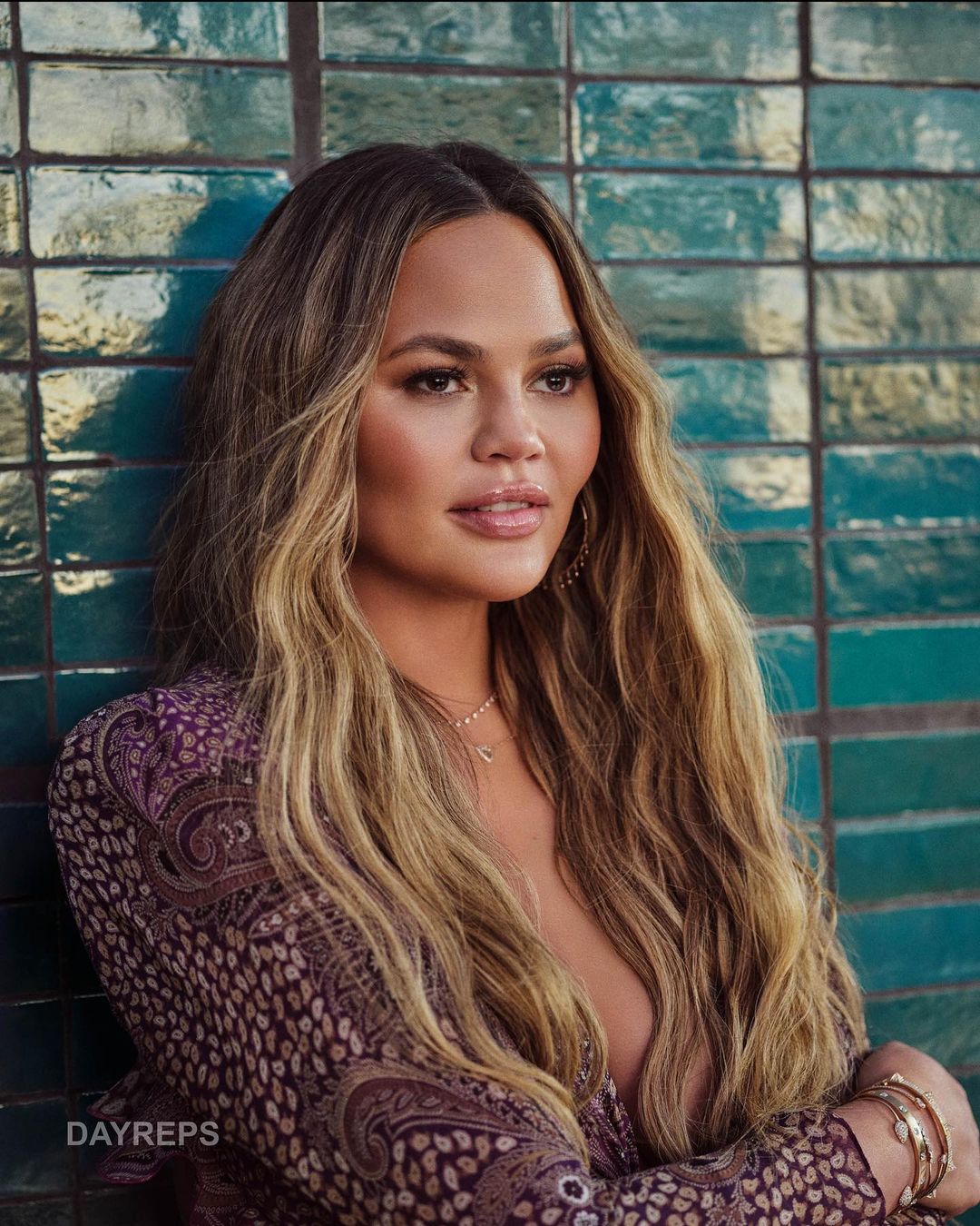 Portrait of Chrissy Teigen posing indoors against a textured greenish-blue tile wall, wearing patterned clothing with gold bracelets and necklace.