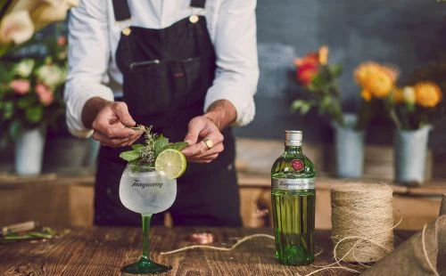 Bartender prepares gin cocktail with lime and herbs, bottle of gin, string, and flowers on wooden bar top with dark background.