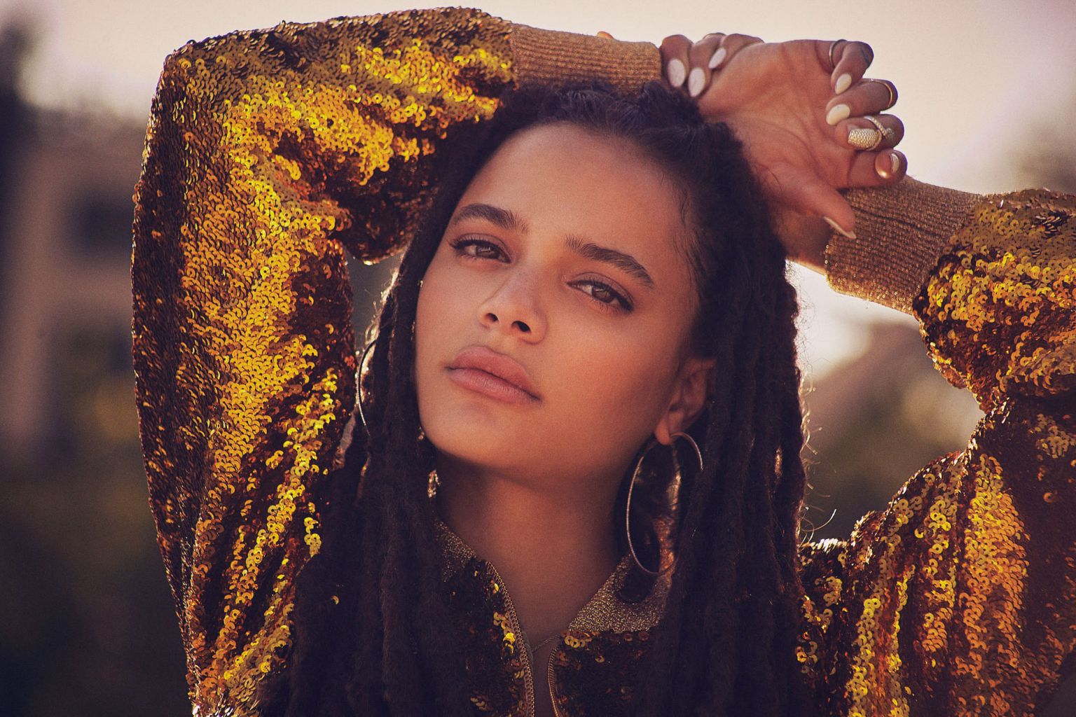 Close-up portrait of a woman with dreadlocks wearing a sequined gold jacket with hands above her head and silver hoop earrings.