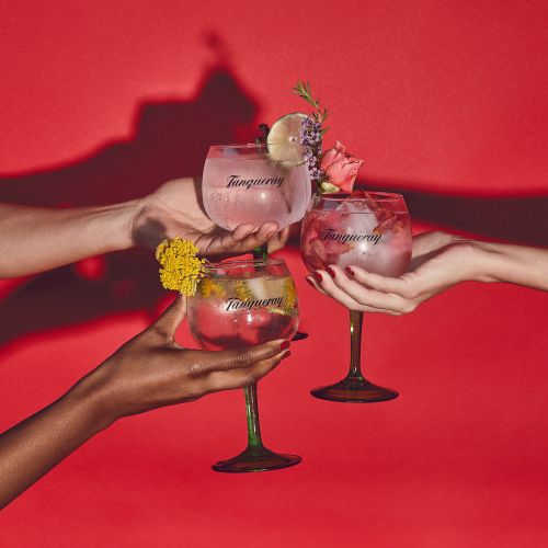 Three cocktails in stemmed glasses are held aloft by three sets of hands against a red background with floral garnishes.