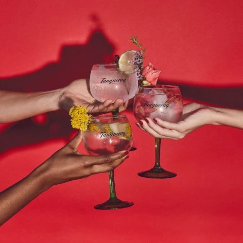 Three cocktails in stemmed glasses are held aloft by three sets of hands against a red background with floral garnishes.