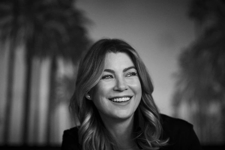 Black and white portrait of a smiling woman with wavy hair wearing a dark blazer against a blurred palm tree backdrop.