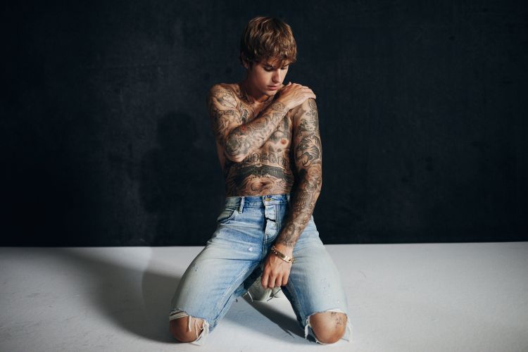 Shirtless man with tattoos kneeling in ripped jeans against a dark backdrop, illuminated by soft light, showcasing his physique.