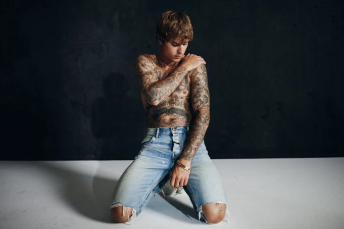 Shirtless man with tattoos kneeling in ripped jeans against a dark backdrop, illuminated by soft light, showcasing his physique.