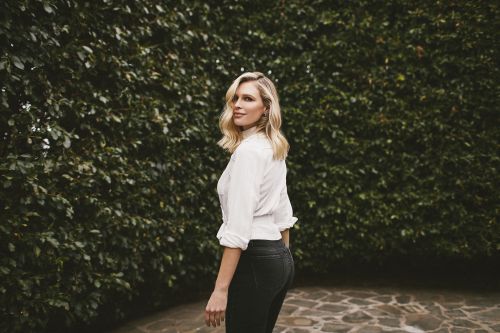 Blonde woman in white shirt and black pants poses in front of a green hedge on a stone pathway in an outdoor setting.