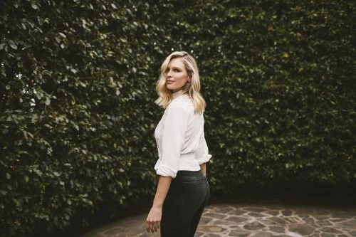Blonde woman in white shirt and black pants poses in front of a green hedge on a stone pathway in an outdoor setting.