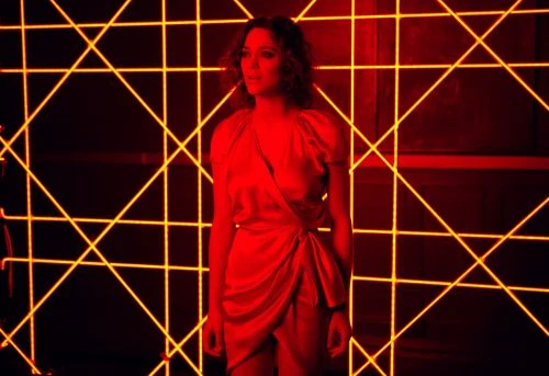 An adult female stands in front of a wall with a geometric laser light display, illuminated with a red glow.