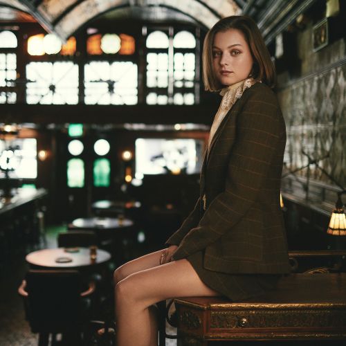 Stylish woman in tweed suit sits on table in bar with vintage design elements, warm lighting, relaxed pose, medium shot indoors.