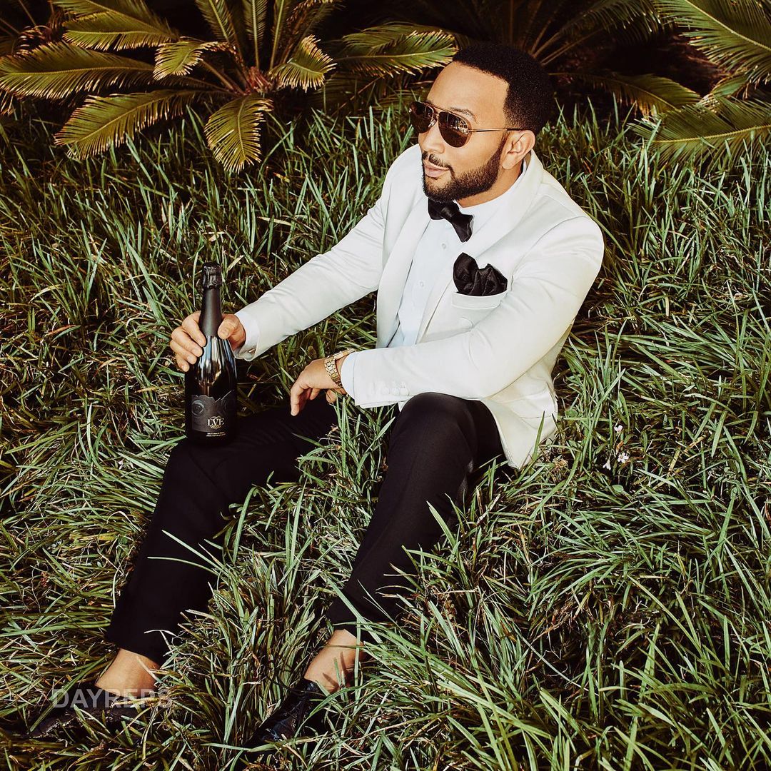 Adult male wearing sunglasses and a suit, sitting in tall grass while holding a bottle, with leafy green plants in the background.
