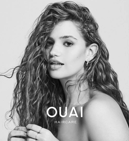 Black-and-white portrait of a smiling woman with long curly hair, safety pin earring, hand rings, and OUAI haircare text overlay.