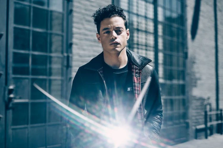 Medium shot of actor Rami Malek in a black jacket and shirt, posing against an urban, gated building backdrop.